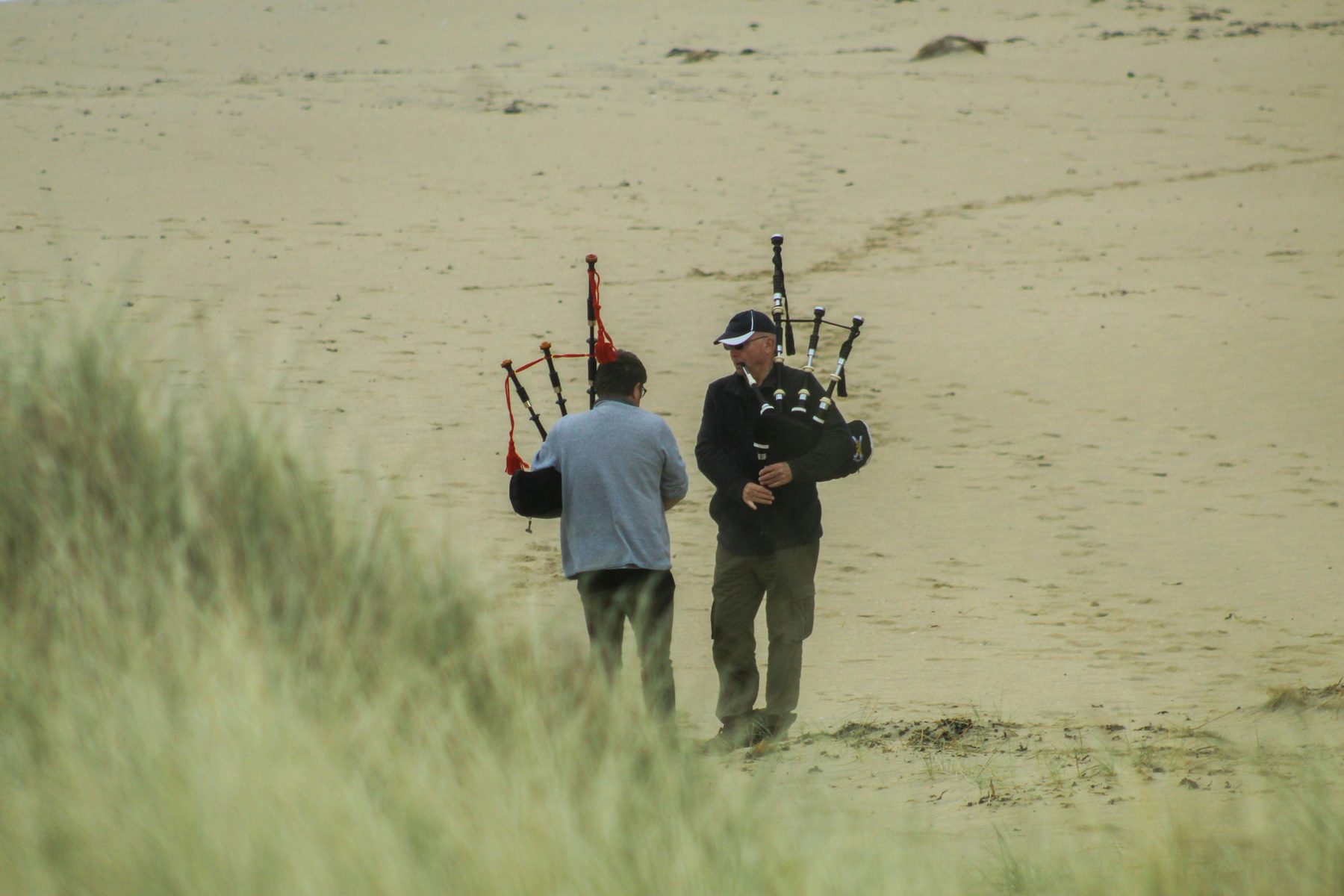 Two figures on a windswept sandy dune with beachgrass in the foreground, one in a shooting jacket and cap playing a full set of Great Highland bagpipes