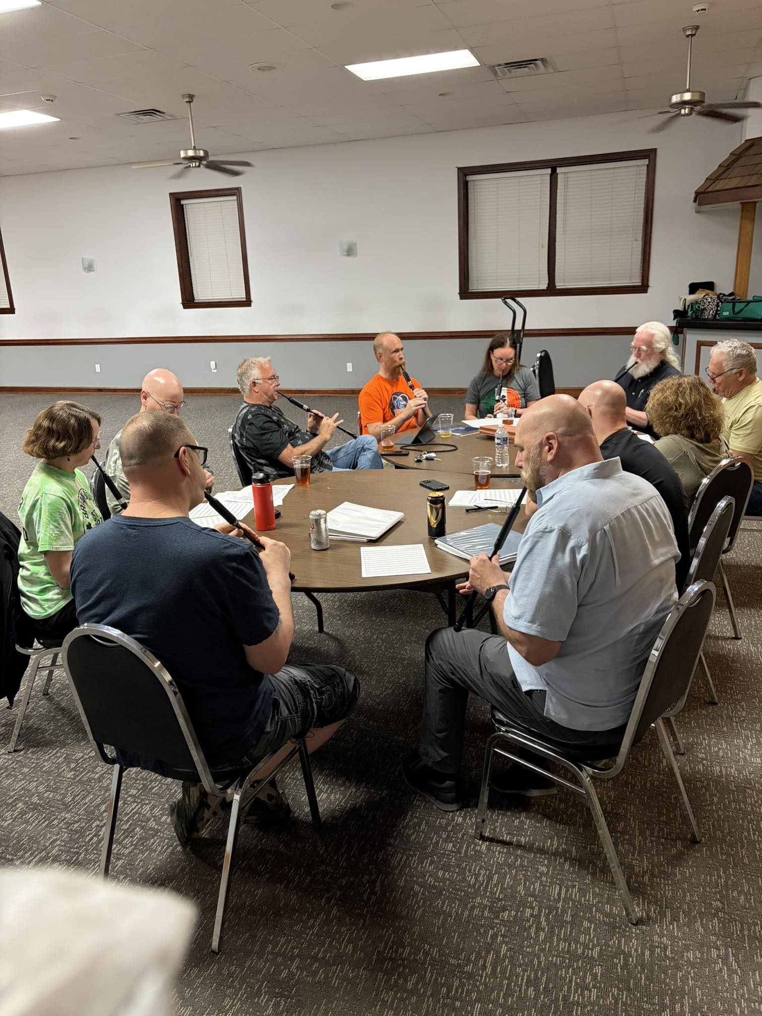 A pipe band practice session in a community-hall setting. About ten people of mixed ages are seated around a round table, most holding practice chanters, with water bottles and mugs on the table between them.
