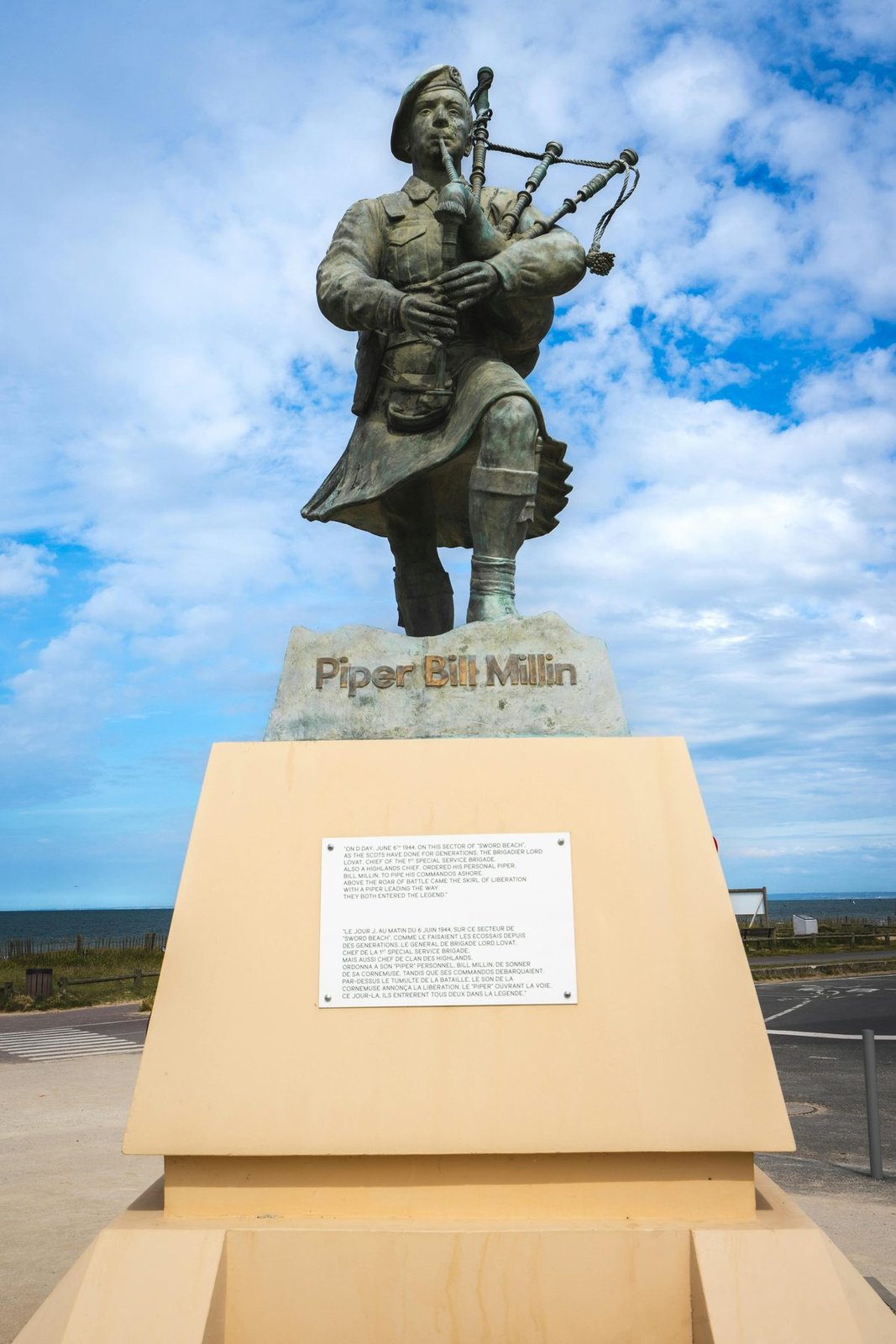 A bronze statue of Piper Bill Millin in full Highland uniform, playing bagpipes, mounted on a stone plinth labeled 'Piper Bill Millin'. The memorial stands near Sword Beach, Normandy, with the sea and sky visible in the background.